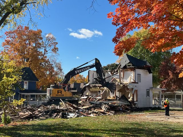 unnamed Photo of a beautiful, crisp fall day in Wisconsin. A demolition digger is pictured tearing down a white house. Fall foliage frames the photo.